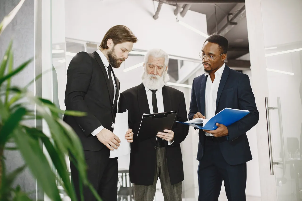 Three people in business dress standing in an office, one holding a blue folder