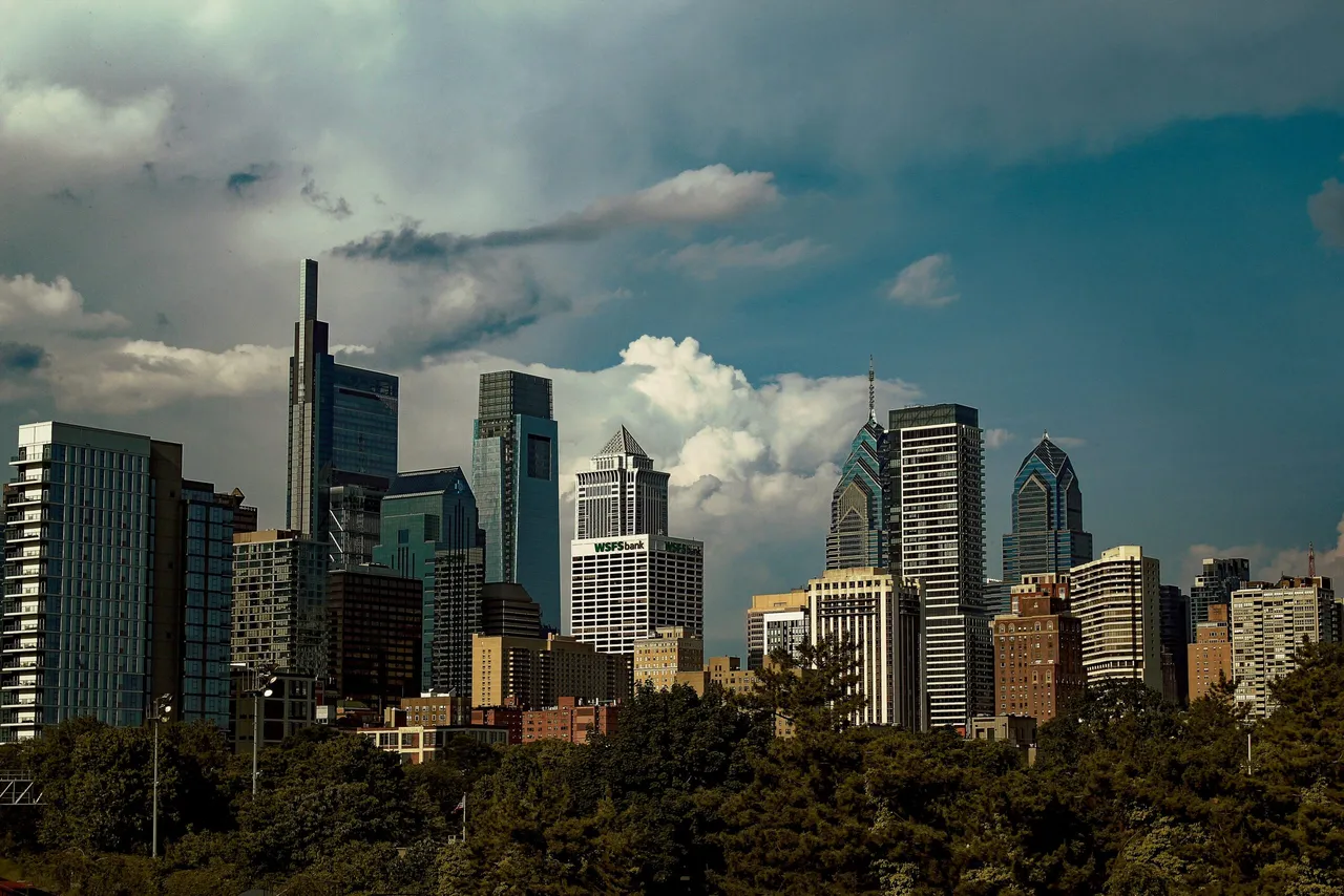 City skyline with tall office buildings and green trees in foreground under blue sky with white clouds