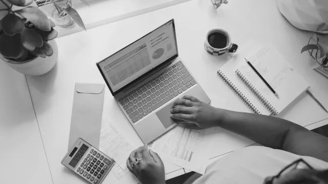 Person working at desk with laptop, coffee cup, notebook and calculator in monochrome