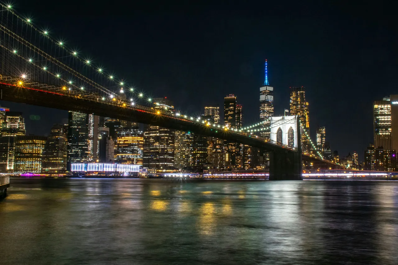Illuminated suspension bridge spanning a river with lit skyscrapers and cityscape at night
