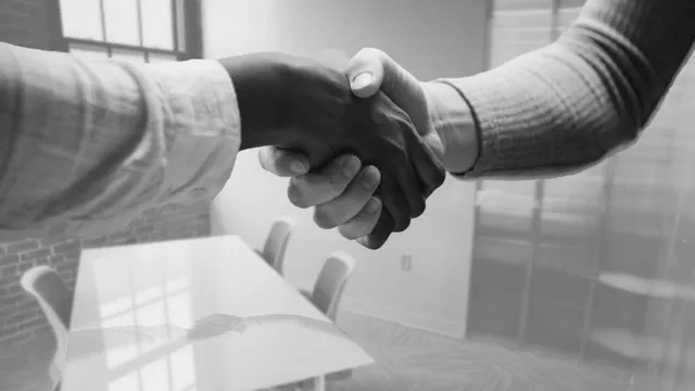 Two people in business attire shaking hands across a conference table in an office setting