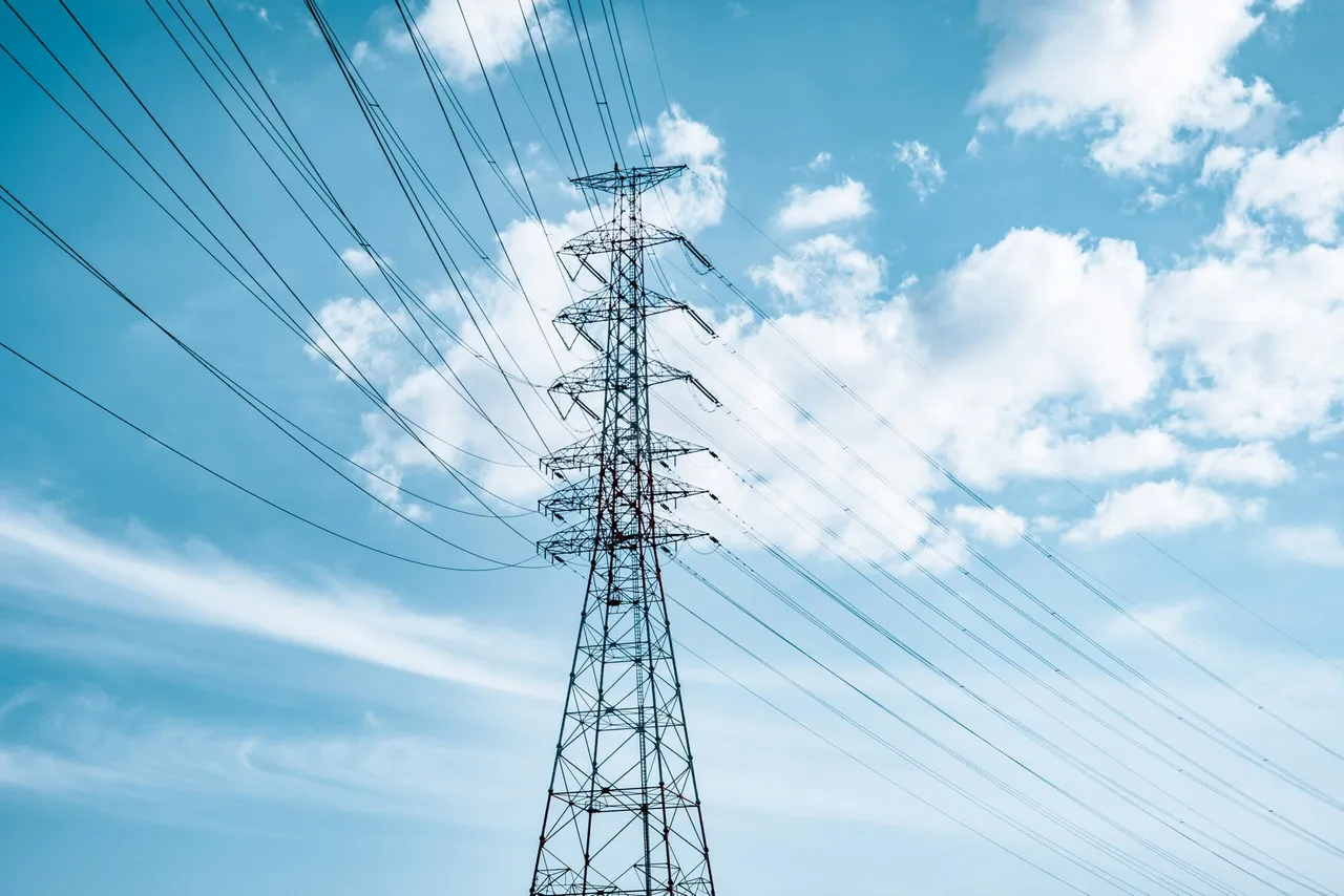 Electricity transmission tower with multiple power lines extending outward against a blue sky with white clouds