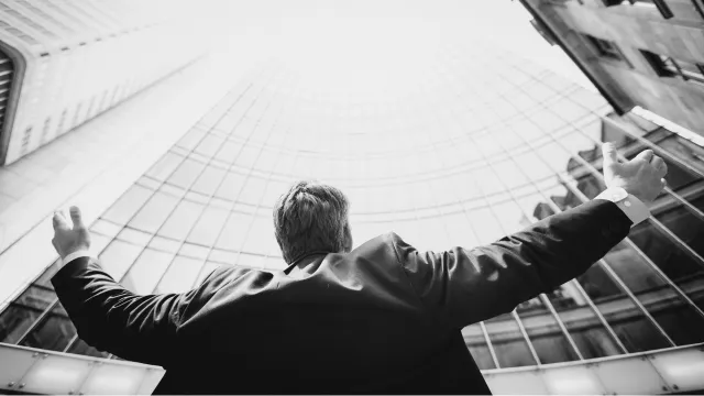 Person with arms raised looking up at modern glass building exterior from below