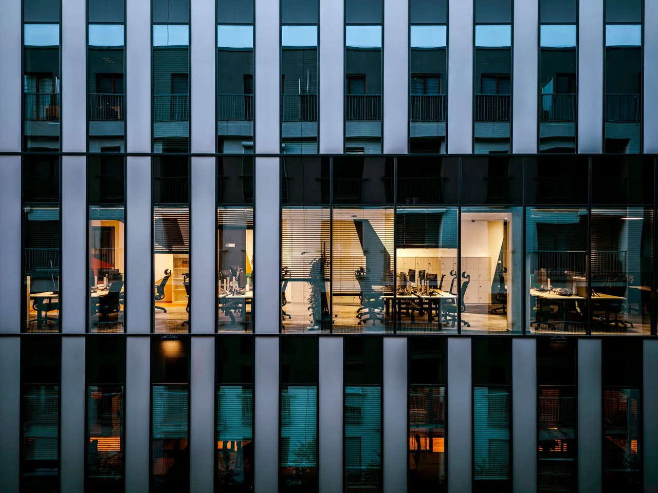 Illuminated office spaces with desks and chairs visible through large windows of a modern building facade at dusk